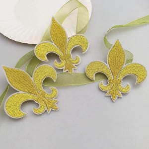 Pode incluir: Três emblemas bordados de flor-de-lis em fio dourado com bordas brancas, dispostos sobre uma fita verde-clara translúcida. Os emblemas apresentam uma textura de fio densa e metálica com um design clássico de três pétalas.