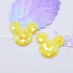 May include: Two yellow mouse-shaped patches covered in small, reflective, circular sequins. Each patch features a rounded head and two circular ears, creating a distinct silhouette. The sequins are arranged in a dense, overlapping pattern.