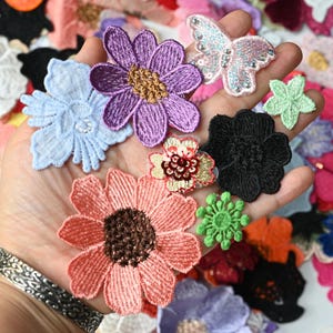 May include: A collection of assorted embroidered fabric patches featuring various floral shapes and a butterfly. The patches include colours such as purple, light blue, peach, black, and green, with some designs incorporating sequins and metallic thread accents.