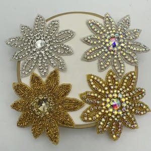 May include: Four flower-shaped patches arranged in a square. Two are silver-toned with clear rhinestones, and two are gold-toned with matching rhinestones. The top right and bottom right flowers feature iridescent, multi-coloured centre stones.