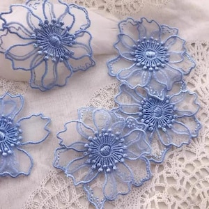 May include: Four light blue floral appliqués featuring intricate embroidery and sheer petal outlines. Each flower has a dense, circular stitched centre with radiating thread details, creating a delicate, layered appearance for these decorative pieces.
