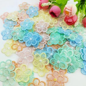 May include: A collection of small, five-petalled flower appliques in pastel shades of blue, yellow, pink, and green. Each piece features intricate, sheer embroidery with a scalloped edge and a central stitched detail, scattered across the frame.