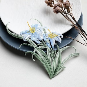 May include: An embroidered patch featuring two light blue lily flowers with yellow centers and detailed green stems and leaves. The embroidery uses varied shades of green thread to create depth in the foliage and light blue thread for the petals.