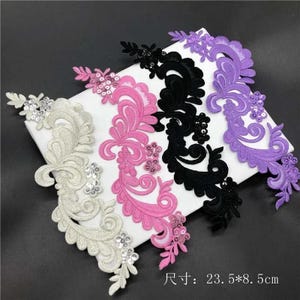 May include: Four embroidered floral appliques in white, pink, black, and purple, each featuring intricate swirling patterns and sequin accents. The appliques measure 23.5 cm by 8.5 cm and are arranged diagonally on a white rectangular surface.