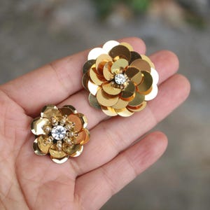 Peut inclure: Deux ornements circulaires en forme de fleur composés de paillettes dorées métalliques. La plus grande pièce présente des pétales superposés avec un strass transparent au centre, tandis que la plus petite possède des perles entourant un strass.