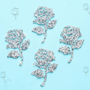 May include: Three rose-shaped iron-on patches covered in clear, sparkling rhinestones. Each patch features a detailed flower head with a stem and two leaves, all constructed from faceted crystals that reflect light against a light blue background.