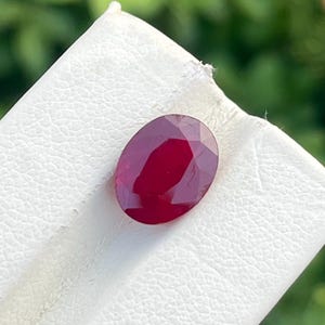 May include: A deep red, oval-cut gemstone with a faceted surface, resting on a white textured display card. The stone exhibits a rich, saturated crimson hue and clear geometric facets that reflect light across its polished, symmetrical surface.