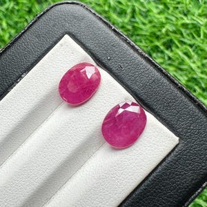 Puede incluir: Dos rubíes rosas facetados de forma ovalada expuestos sobre un inserto de terciopelo blanco dentro de un estuche negro. Las piedras preciosas muestran una calidad translúcida con inclusiones internas visibles y una superficie pulida.