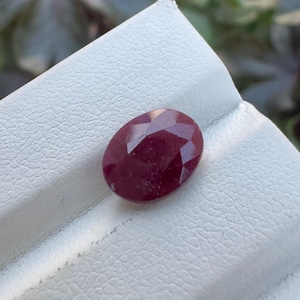 May include: An oval-shaped, faceted deep red ruby gemstone with a polished surface. The stone displays a rich, saturated crimson hue and is set against a textured, off-white display surface with a diagonal crease running across the centre.
