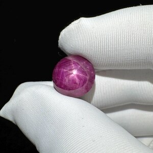 May include: A round, cabochon-cut gemstone with a deep magenta-pink hue. The stone displays a distinct six-rayed star pattern across its surface, characteristic of asterism, with a smooth, polished finish and a translucent appearance.