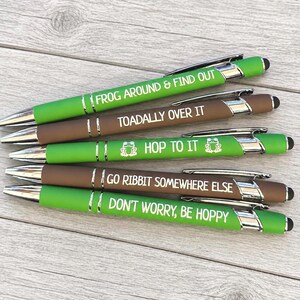May include: A set of five ballpoint pens with stylus tips, featuring green and brown matte barrels. Each pen displays white text: FROG AROUND & FIND OUT, TOADALLY OVER IT, HOP TO IT with frog icons, GO RIBBIT SOMEWHERE ELSE, and DON'T WORRY, BE HOPPY.