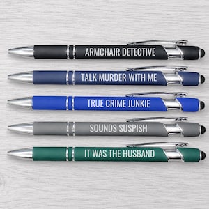 May include: Five retractable ballpoint pens with silver-tone clips and tips. The pens are black, navy, blue, gray, and green, each featuring white text: ARMCHAIR DETECTIVE, TALK MURDER WITH ME, TRUE CRIME JUNKIE, SOUNDS SUSPISH, and IT WAS THE HUSBAND.