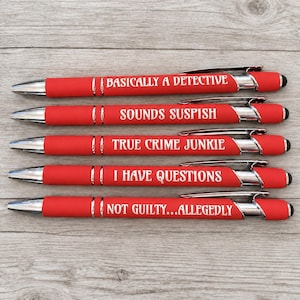May include: Five red retractable ballpoint pens with silver accents and black stylus tips. Each pen features white text: BASICALLY A DETECTIVE, SOUNDS SUSPISH, TRUE CRIME JUNKIE, I HAVE QUESTIONS, and NOT GUILTY...ALLEGEDLY, arranged in a vertical stack.