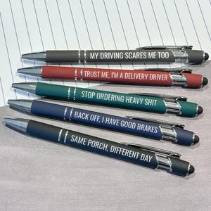 May include: A set of five retractable ballpoint pens with stylus tips, featuring chrome accents and white text. The pens are colored charcoal, red, teal, navy, and dark blue, each printed with humorous phrases related to delivery driving.