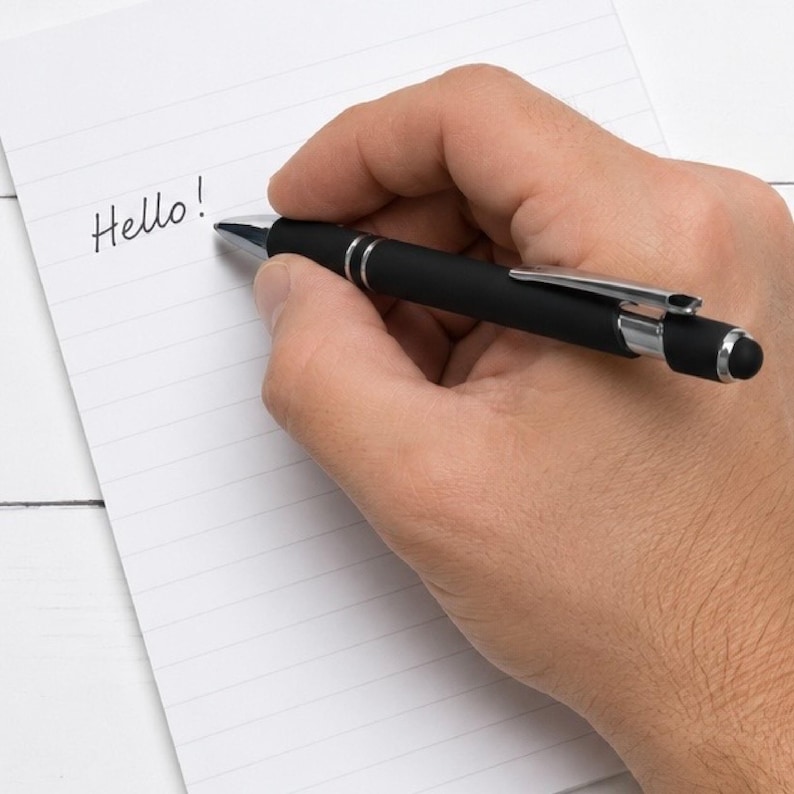 May include: A matte black ballpoint pen with a silver clip and a black stylus tip at the end, positioned on a white lined notepad with the word Hello written in black ink. The pen features a sleek design with silver accents near the grip area.