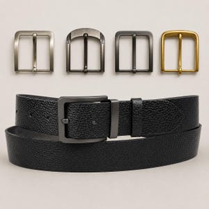 Op de afbeelding: Een zwarte riem met een getextureerd oppervlak en een donkergrijze gesp wordt getoond onder vier losse riemgespen in zilver, geborsteld gunmetal, matzwart en antiek goud. De riem heeft een generfde textuur en een enkele bijpassende lus.
