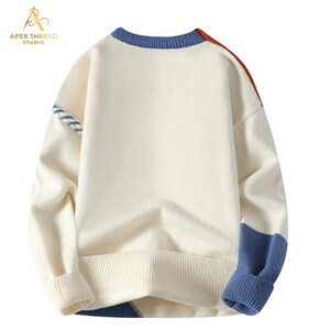 May include: A cream-colored long-sleeve knit sweater featuring a blue ribbed crew neckline, cuffs, and hem. The sweater includes color-blocked accents in blue and red on the shoulders and lower sections, with a blue and white striped detail on one sleeve.