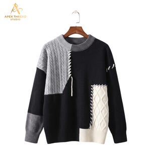 May include: A long-sleeve color-blocked sweater featuring black, gray, and white panels. The garment includes a gray cable-knit section on the upper right, a white diamond-patterned knit panel on the lower right, and white decorative lacing details.
