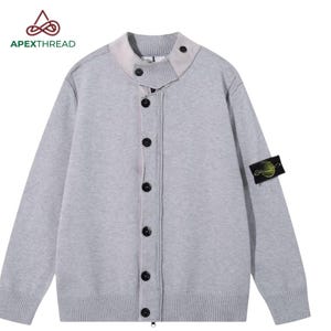 Könnte beinhalten: Eine hellgraue Strickjacke mit hohem Kragen und Knopfverschluss, einer durchgehenden Knopfleiste mit dunklen Knöpfen sowie einem schwarzen rechteckigen Aufnäher mit kreisförmigem Logoemblem am linken Ärmel des Kleidungsstücks.