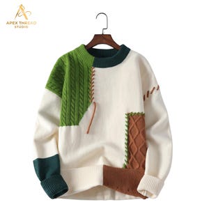 May include: A long-sleeve crewneck sweater featuring a color-blocked design in cream, forest green, and brown. The garment includes sections of cable-knit texture, visible whip-stitch detailing in brown and green thread, and a ribbed collar and cuffs.