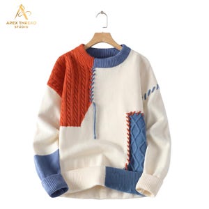Puede incluir: Un suéter de manga larga con cuello redondo que presenta un diseño de bloques de color en crema, naranja quemado y azul. La prenda incluye paneles de punto trenzado, puños y dobladillo acanalados, y detalles de costura decorativa.