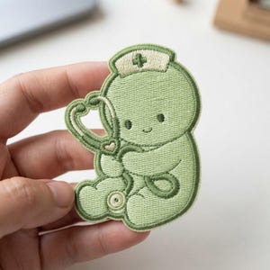 May include: A light green embroidered patch featuring a cartoon character wearing a nurse cap with a cross symbol. The character is holding a heart-shaped stethoscope. The design uses shades of sage green and cream thread with a dark green border.