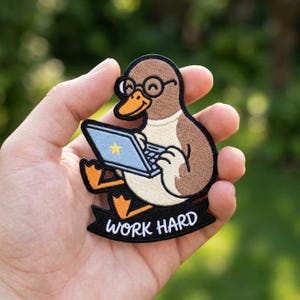 May include: An embroidered patch featuring a brown and cream-colored duck wearing round black glasses. The duck is typing on a light blue laptop with a yellow star icon. Below the duck is a black banner with the white text WORK HARD.