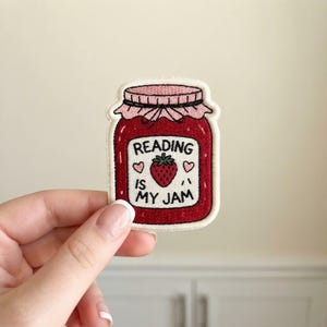 May include: An embroidered patch shaped like a jar of red strawberry jam with a light pink lid. The label features a central strawberry illustration, two small pink hearts, and the black text Reading Is My Jam, all outlined in cream.