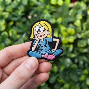 May include: An embroidered patch featuring a cartoon character with blonde hair, wearing a light blue scrub top and pants with a stethoscope around the neck. The character is sitting with legs crossed, resting a chin on a hand, wearing pink shoes.