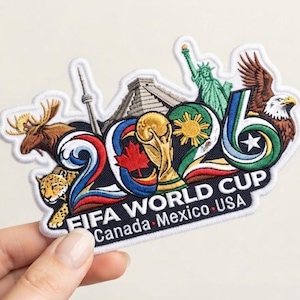 May include: An embroidered patch featuring the text FIFA WORLD CUP CANADA MEXICO USA and the number 2026. The design includes a moose, a jaguar, the CN Tower, a pyramid, the Statue of Liberty, a bald eagle, and the FIFA World Cup trophy.