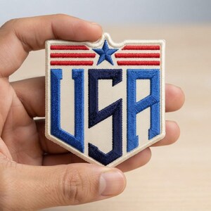 May include: A shield-shaped embroidered patch with a cream-colored background. The design features the letters USA in bold blue and dark blue stitching, topped with a blue star and three horizontal red stripes on each side of the star.