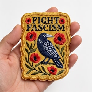 May include: A rectangular embroidered patch with a scalloped edge featuring a yellow background, a dark blue crow, and red poppy flowers. The text FIGHT FASCISM is embroidered in dark blue capital letters at the top of the patch design.