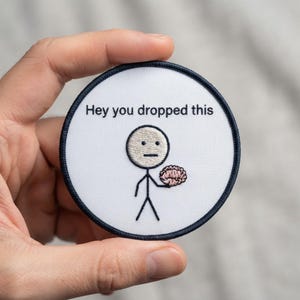 May include: A circular embroidered patch with a white background and a dark blue border. The patch features a black stick figure holding a pink brain, with the black text "Hey you dropped this" printed in a simple sans-serif font above the figure.