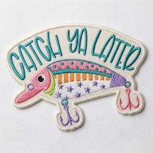 May include: An embroidered patch featuring a colorful fishing lure with a pink head, orange and white striped body, and purple star patterns. The text "CATCH YA LATER" is stitched in teal above the lure, which includes two purple fishing hooks.