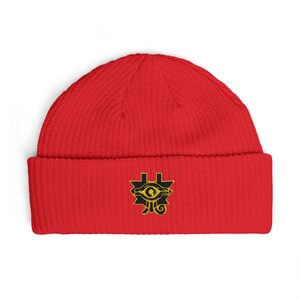 Puede incluir: Un gorro de punto rojo brillante con un dobladillo plegado. Centrado en el dobladillo hay un parche bordado que muestra un gráfico negro y amarillo del Ojo de Horus, un antiguo símbolo egipcio, con líneas geométricas y una pupila central.