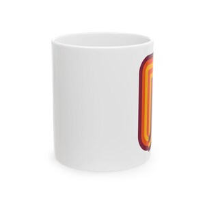 May include: A white ceramic mug featuring a vertical graphic design on the side. The graphic consists of four concentric rounded rectangular shapes in shades of dark maroon, bright orange, golden yellow, and light yellow, creating a layered effect.