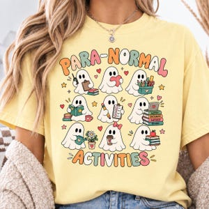 May include: A pale yellow t-shirt featuring a graphic print with the text PARA-NORMAL ACTIVITIES. The design displays nine white ghosts engaged in various tasks, including holding books, a clipboard, a pencil, a coffee cup, and a potted flower.