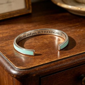 Könnte beinhalten: Ein silberfarbenes Armband mit einer glatten, helltürkisfarbenen Emaileinlage auf der Außenseite. Die Innenseite ist mit dem schwarzen Text "This too shall pass, BUT FUCKING HELL" in einer klaren Serifenschrift graviert.