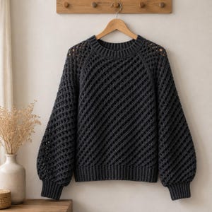 May include: A dark charcoal grey long-sleeve jumper featuring a diagonal open-knit crochet pattern, a round ribbed neckline, and ribbed cuffs. The garment has a relaxed fit with dropped shoulders and a solid ribbed hem at the bottom edge.