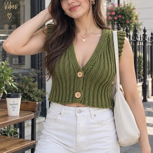 Peut inclure: Un gilet sans manches en tricot côtelé vert olive avec un décolleté en V profond, des détails à volants sur les épaules et deux gros boutons en bois clair sur le devant. Le vêtement est associé à un pantalon en jean blanc et un sac à épaule blanc.