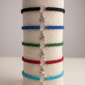 Può includere: Un set di cinque braccialetti in corda intrecciata nei colori nero, blu reale, verde bosco, rosso e turchese. Ogni braccialetto presenta un anello centrale in metallo argentato con un piccolo ciondolo a forma di stella argentata.