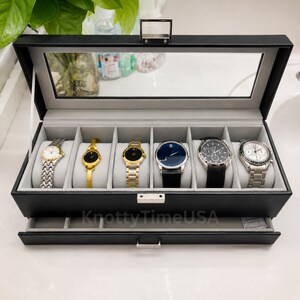 May include: A black rectangular watch display case with a clear glass lid and a silver metal latch. The interior is lined with light gray fabric and features six individual compartments, each containing a wristwatch, plus a pull-out drawer below.