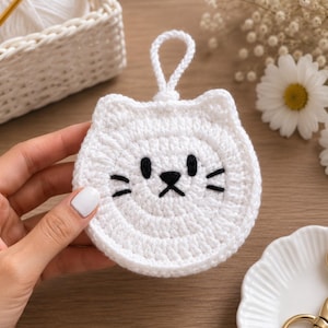 May include: A round, white crocheted cat face with a small loop at the top for hanging. The cat features embroidered black yarn details, including two oval eyes, a small triangular nose, and three whiskers on each side of the face.