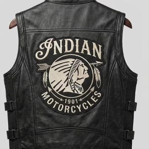 May include: A black leather vest featuring a large embroidered patch on the back. The patch displays the text Indian Motorcycles 1901 and an illustration of a person wearing a feathered headdress. The vest has side adjustment straps and panels.