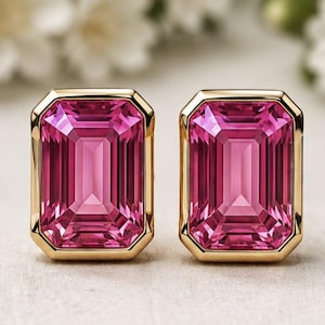 May include: A pair of stud earrings featuring large, rectangular, emerald-cut pink gemstones set in polished yellow gold bezels. The stones exhibit a deep magenta hue with multifaceted surfaces that reflect light, creating a clear, brilliant appearance.