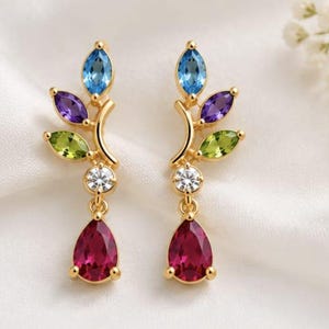 Könnte beinhalten: Ein Paar goldfarbene Ohrringe mit einem zweigartigen Design und marquise-geschliffenen blauen, violetten und grünen Edelsteinen. Jeder Ohrring enthält einen runden, klaren Stein und einen hängenden, birnenförmigen, dunkelrosa Edelstein.