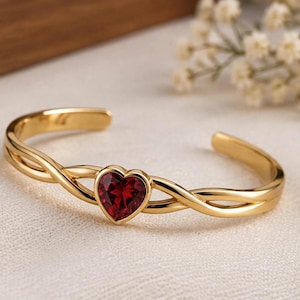 Pode incluir: Uma pulseira tipo bracelete em tom dourado com acabamento polido e uma pedra preciosa vermelha em forma de coração ao centro. A banda consiste em dois fios metálicos entrelaçados que se encontram no engaste do coração, criando um design aberto.
