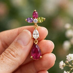 Könnte beinhalten: Ein goldfarbener Ohrhänger mit einem mehrfarbigen Blumenmotiv an der Oberseite, bestehend aus einem zentralen klaren Stein, einem violetten, einem grünen und einem roten Markisenschliff-Stein, gefolgt von einem klaren birnenförmigen Stein und einem roten Tropfen.