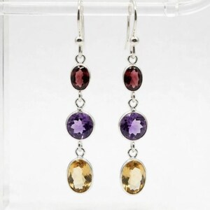 Könnte beinhalten: Ein Paar silberne Ohrhänger mit drei vertikal angeordneten facettierten Edelsteinen. Jeder Ohrring zeigt oben einen dunkelroten ovalen Stein, in der Mitte einen runden violetten Amethyst und unten einen hellgelben ovalen Citrin.