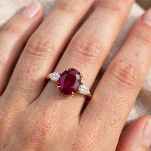 Könnte beinhalten: Ein Goldring mit einem großen, oval geschliffenen, tiefroten Rubin in der Mitte, der von zwei kleineren, tropfenförmig geschliffenen weißen Diamanten flankiert wird. Die Edelsteine sind in einer polierten Goldschiene mit Krappenfassung gefasst.